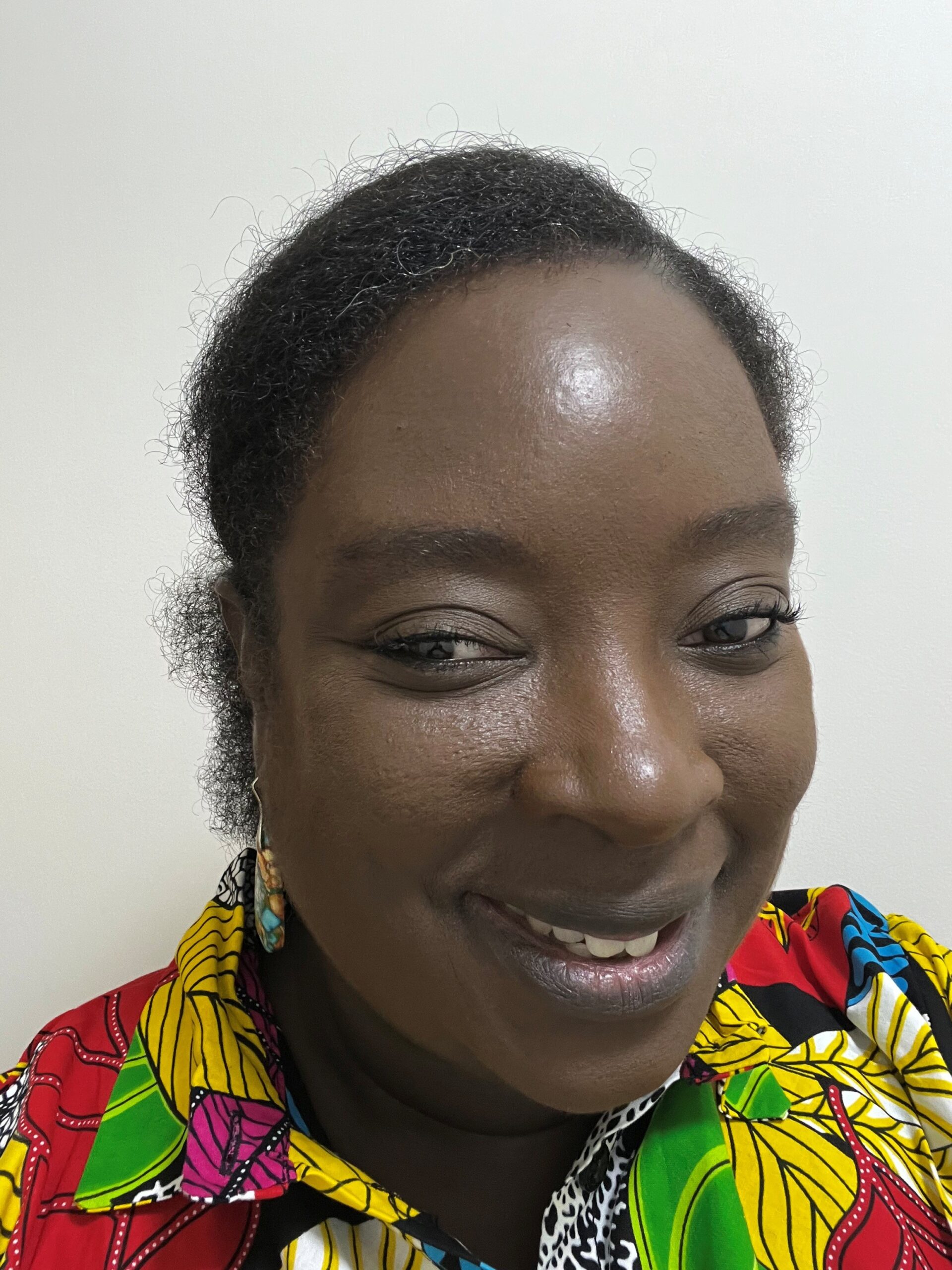 Ruth, a smiling woman with dark skin and short curly hair wearing a colourful patterned shirt with red, yellow, green, and blue elements.