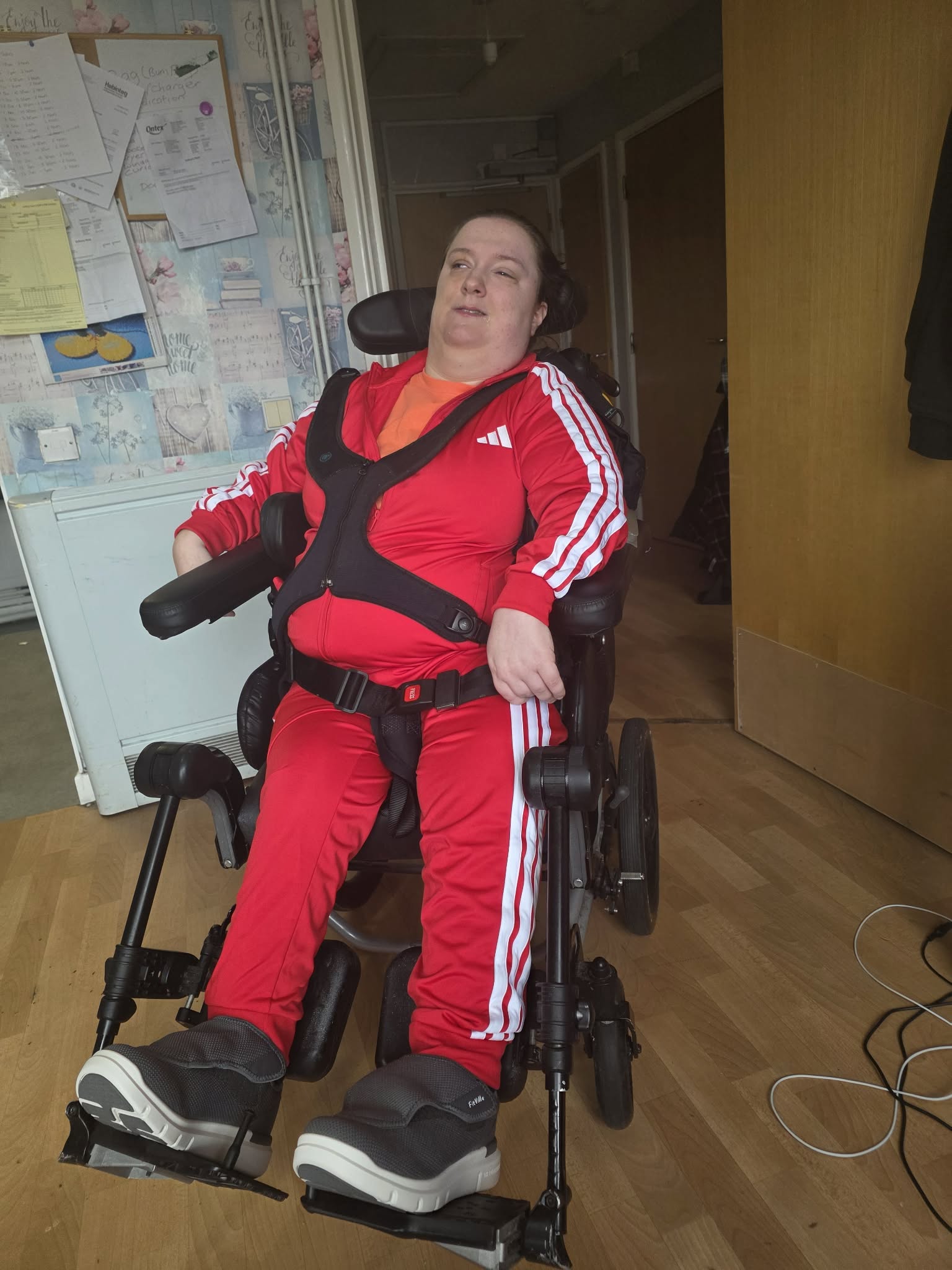 Terri is a person in a wheelchair. She is wearing a red tracksuit with white lines going down the arm and leg.