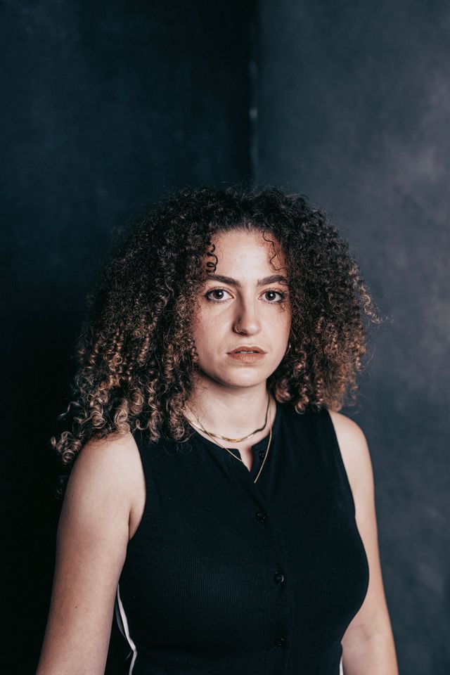 Yiska is a mixed heritage woman in her late twenties with long curly hair, looking at the camera and standing against a dark background.