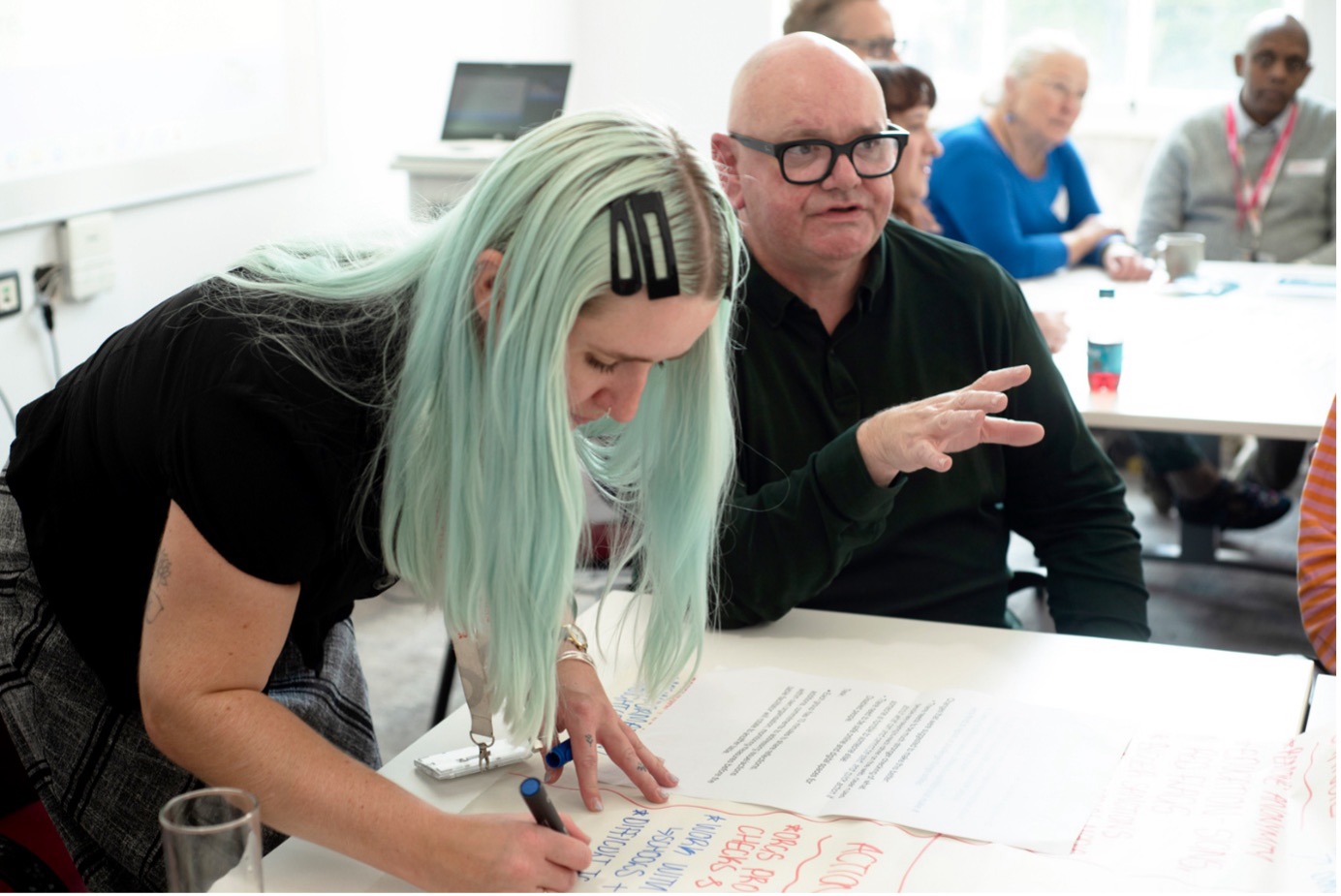 A woman with blue hair writes down comments from the table discussions on a large piece of paper.